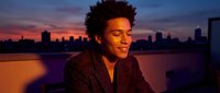 Young African American businessman smiling on rooftop terrace at sunset with city skyline background. Professional portrait for corporate marketing materials.