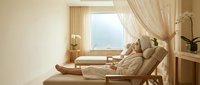 Relaxed woman in bathrobe enjoying spa treatment on wooden lounge chair in bright wellness center with natural lighting and peaceful atmosphere.