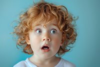 Surprised redhead child with wide eyes and curly ginger hair expressing amazement against blue background. Emotional portrait showing genuine wonder and astonishment.