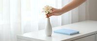 Woman's hand placing white flowers in ceramic vase on desk with blue notebook, bright minimalist workspace interior design and home office decoration.