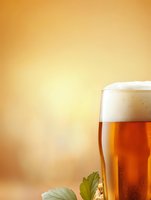 Fresh draft beer with thick white foam head and green hop leaves against warm golden background, condensation droplets on glass surface.