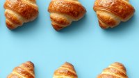 Fresh baked golden croissants arranged in rows on light blue background, creating minimalist pattern composition for food photography and menu design.