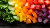 Fresh colorful chopped vegetables arranged in rainbow gradient pattern with lettuce, cucumber, yellow and orange peppers, tomatoes and red onion on dark wood.