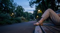 Person relaxing on park bench during twilight hour with warm street lighting and lush green trees creating peaceful evening atmosphere for wellness content.