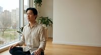 Asian man meditating in modern apartment with city view, practicing mindfulness and wellness in bright living space with plants and wooden floors.
