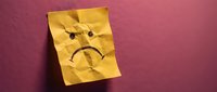 Crumpled yellow sticky note with hand-drawn sad face expression on textured pink wall background for emotional communication and office concepts.