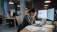 Stressed businesswoman holding head in hands at office desk with laptop and financial charts during late night work session in modern coworking space.