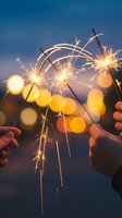 Hands holding lit sparklers against twilight sky with bokeh lights, celebrating festive occasions, New Year's Eve, birthdays, and outdoor nighttime events.
