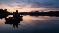 Fisherman in small boat on calm lake at sunset with mountains in background, suitable for outdoor recreation and lifestyle content.