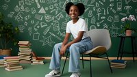Young African American girl student sitting on modern chair in classroom with educational doodles on green chalkboard background for back to school concepts.