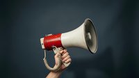 Red and white megaphone held by hand against dark background for communication, announcement, marketing campaigns and promotional activities.