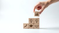 Hand placing wooden cube with growth chart icon completing business success pyramid with lightbulb handshake graduation cap symbols on white background.