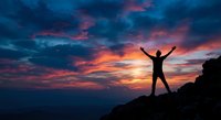 Silhouette of person with raised arms celebrating success on mountain peak against dramatic sunset sky with vibrant orange and blue clouds.