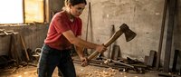 Young Caucasian male carpenter swinging axe in rustic workshop with wooden beams and tools, traditional craftsmanship and woodworking skills in action.