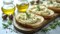 Rustic bread slices topped with creamy white bean spread, fresh rosemary herbs and olive oil on wooden cutting board for healthy Mediterranean appetizer recipes.