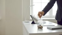 Business professional adding coins to glass jar on modern office desk with laptop and documents for savings and financial planning goals.