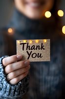 Female hand holding glitter thank you card with bokeh lights in background, wearing cozy knit sweater. Intimate gratitude moment captured in warm atmospheric lighting.