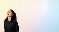 Young woman with long dark curly hair and medium skin tone wearing black shirt, looking upward with positive expression against soft pastel gradient background.