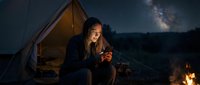 Woman with long dark hair, wearing dark clothing, sitting by campfire at night under starry sky with Milky Way visible, camping tent glowing in background, outdoor adventure and nighttime wilderness scene.