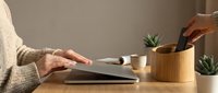 Cozy home office workspace with woman in knit sweater opening laptop on wooden desk with plants and coffee cup.