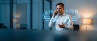 Mature male professional in light blue shirt talking on phone with digital icons floating around him in modern office setting with warm lighting.