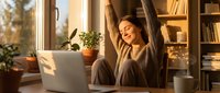 Young Caucasian woman stretching with eyes closed at home desk near laptop, enjoying warm sunlight, cozy remote work lifestyle concept.