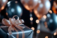 Elegant gift box wrapped in gray paper with peach satin ribbon bow against blurred festive background with balloons and soft bokeh lights.