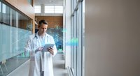 Professional male doctor in white coat using digital tablet in modern hospital corridor with glass walls and natural lighting for healthcare technology.