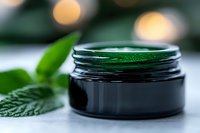 Natural organic green cosmetic cream in dark glass jar with fresh mint leaves and soft bokeh lights on white background, skincare concept.