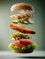 Deconstructed crispy chicken burger with fresh ingredients floating in air showing all layers including sesame bun, lettuce, tomato, pickles and breaded fillet.