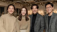 Four young Asian professionals smiling at camera in modern gallery space with warm lighting and contemporary art displays in background.