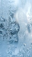 Delicate ice crystals forming intricate frost patterns on glass surface with soft blue tones and bokeh lights creating magical winter atmosphere.