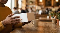 Young adult male with dark skin tone holding blank white card at wooden table in modern cafe setting with blurred background, suitable for business, marketing, and hospitality concepts.