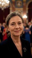 Mature Caucasian woman with gray hair in elegant black attire at formal event in ornate ballroom with chandelier and classical architecture background.