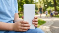 Child holding blank white mobile device outdoors in park with blurred background, suitable for app mockups and technology advertising.