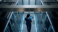 IT technician with tablet walking through modern data center server room with glowing blue lights for technology and cloud computing projects.