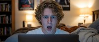 Young man with curly blonde hair and fair complexion displaying shocked expression while using laptop at night in cozy bedroom with warm lighting and bookshelves.