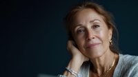 Mature woman with gentle smile against dark background, hand thoughtfully resting on neck. Soft lighting highlights natural beauty and contemplative expression.