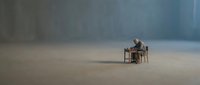 Middle-aged man sitting alone at wooden desk in vast empty concrete space, contemplating solitude and isolation in minimalist environment.