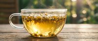 Herbal tea in transparent glass cup with dried herbs floating, warm golden liquid on wooden table with blurred green garden background for wellness concepts.