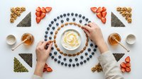 Hands arranging healthy breakfast bowl with yogurt, blueberries and strawberries in geometric pattern. Nutritious morning meal preparation from above.
