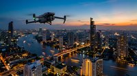 Drone flying over illuminated city skyline at dusk with river winding through skyscrapers, vibrant sunset sky in background.