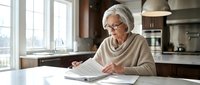 Senior Caucasian woman with glasses reading documents at kitchen table, reviewing paperwork in bright modern home interior for financial planning.
