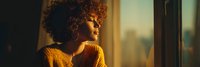 Young African American woman with curly hair wearing orange sweater looking thoughtfully out window during golden hour sunset light for lifestyle content.
