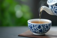 Pouring hot tea from traditional blue and white porcelain teapot into matching teacup on wooden surface against blurred green background.