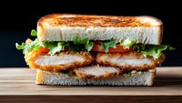 Crispy chicken sandwich with fresh lettuce and tomato on toasted white bread, served on wooden surface against dark background. Close up view shows layered ingredients.