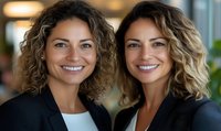 Two professional Hispanic women with curly ombre hair and bright smiles wearing business attire pose confidently in office setting, representing leadership and success.