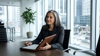Mature woman with gray hair and medium skin tone in professional business attire working at desk with tablet in modern high-rise office with city skyline view.