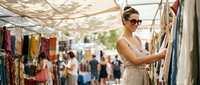 Woman in sunglasses browsing clothing at outdoor market stall, shopping for fashion apparel among vendors and customers on sunny day.