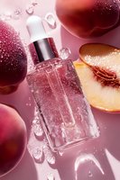 Glass dropper bottle with fresh peach fruits and water drops on pink background, cosmetic skincare product photography for beauty industry advertising.